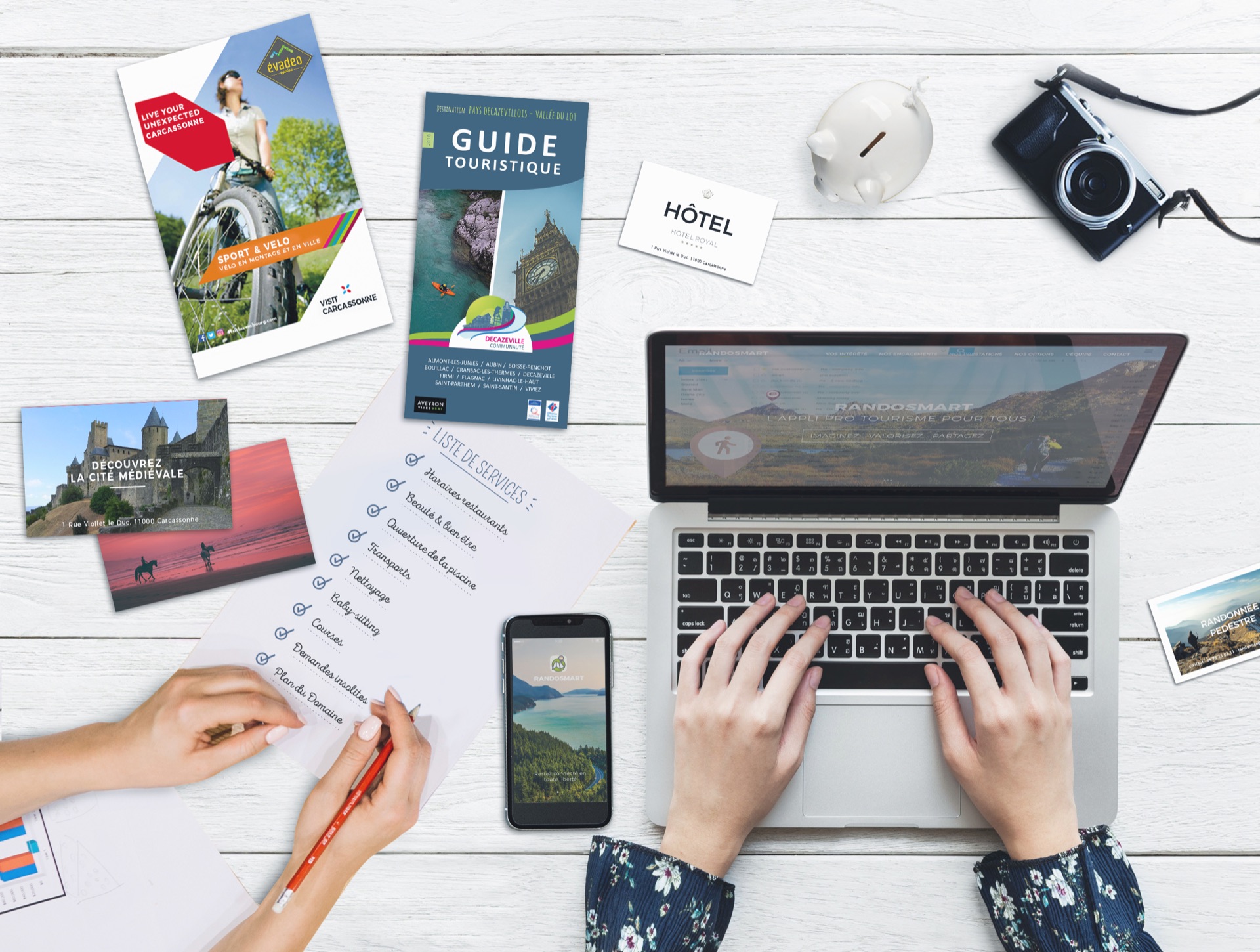 Un bureau vu de haut : brochures touristiques, smartphone affichant Randosmart et ordinateur portable ouvert sur l'interface administrateur.
